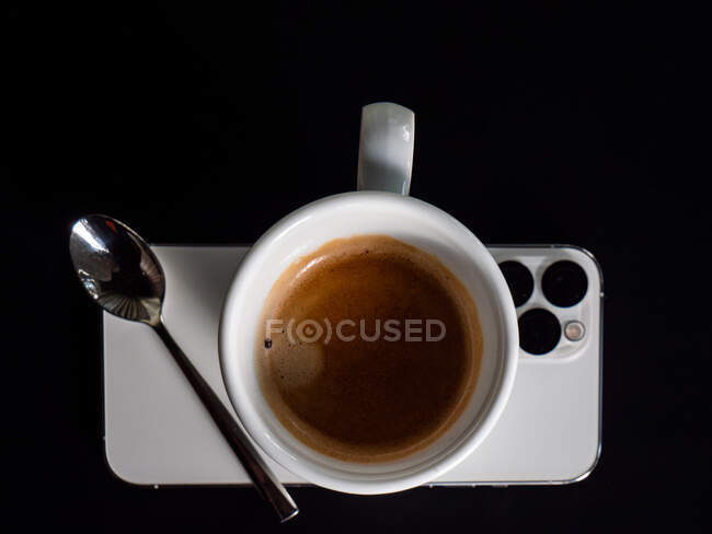 Vista superior de la taza de café expreso y cuchara de metal colocado en el teléfono móvil moderno sobre fondo negro - foto de stock