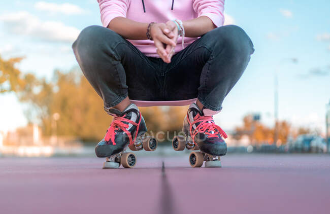 Cropped unrecognizable female wearing light pink hoodie and black jeans and roller skates with neon pink shoelaces squatting down in skate park — Stock Photo