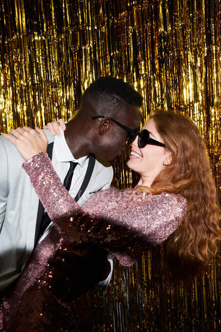 Content young multiethnic couple in modern sunglasses embracing in moment of kiss against tinsel during New Years Eve celebration — Stock Photo