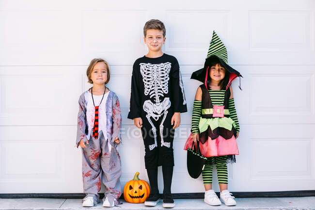 Cuerpo completo de grupo de niños pequeños vestidos con varios disfraces de Halloween con linterna Jack O tallada de pie cerca de la pared blanca en la calle - foto de stock