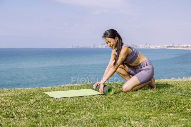 Задоволена молода азіатка в спортивному одязі викидає кору йоги на узбережжі трави проти безконечного океану на сонці. — стокове фото