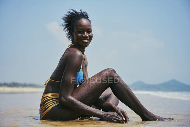 Веселая молодая этническая женщина в купальниках с булочкой из афро-волос, смотрящая в сторону, сидя на берегу океана под голубым небом — стоковое фото