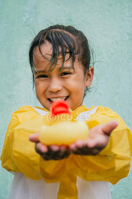 Cheerful ethnic kid in slicker with wet hair and rubber duck looking at camera on pastel background — Stock Photo