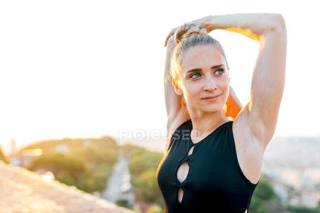 Ajuste femenino en ropa deportiva que estira los brazos durante la práctica de yoga en la azotea del edificio al atardecer - foto de stock