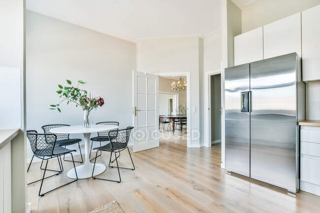 Interno di cucina luminosa con armadi bianchi e frigo con zona pranzo arredata con tavolo e sedie in metallo nero — Foto stock