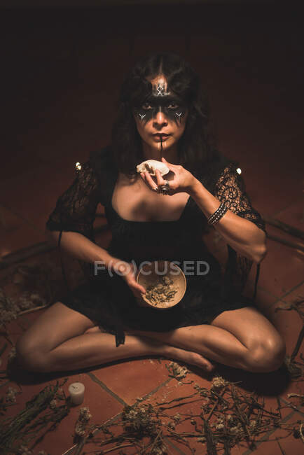 From above of enigmatic witch in black dress sitting in circle made of branches and burning candles during spiritual ritual in dark room holding skull and looking at camera — Stock Photo