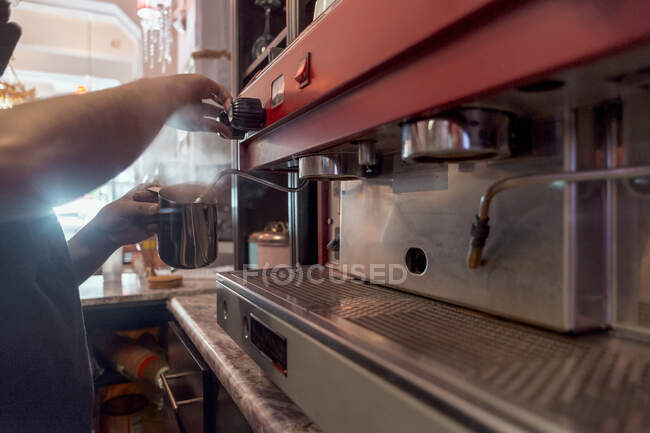Crop anonymous cafeteria employee against professional coffee maker with metal pitcher in cafeteria kitchen on blurred background — Stock Photo