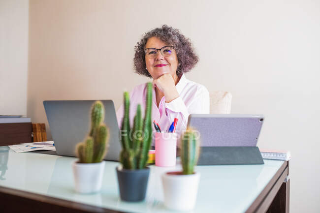 Allegro anziana donna d'affari in occhiali seduto a tavola con netbook e mouse mentre guarda la fotocamera su sfondo chiaro — Foto stock