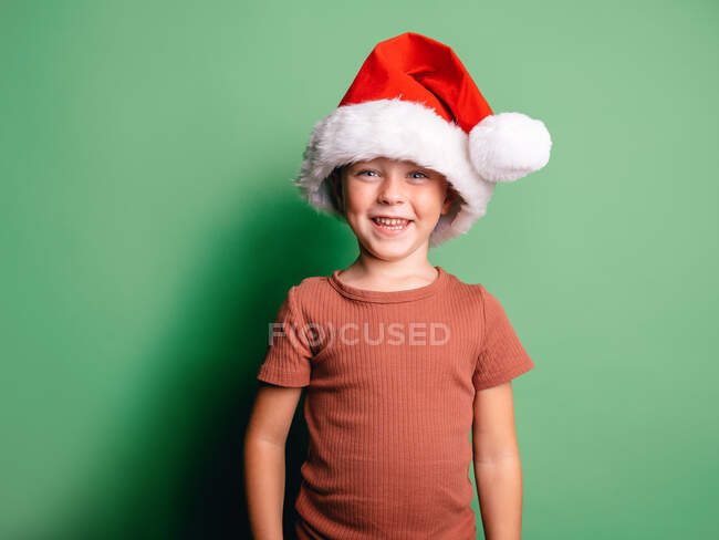 Positivo niño pequeño con el sombrero de Santa rojo sonriendo ampliamente contra el fondo verde y mirando a la cámara - foto de stock