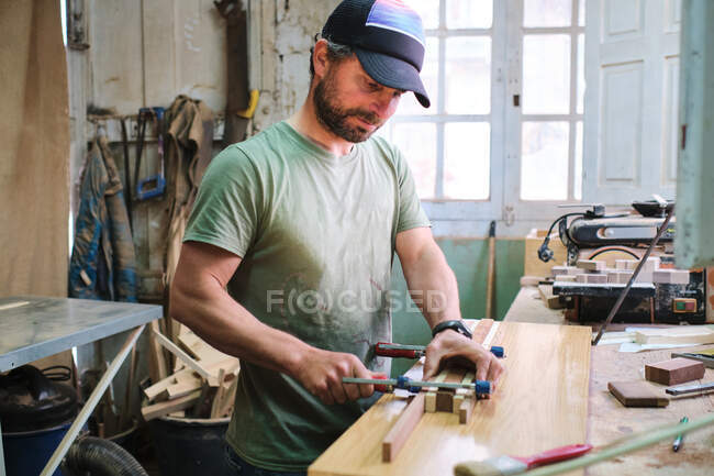 Aufmerksame bärtige männliche Holzarbeiter in Mütze Kleben und Klemmen von Holzblöcken während der Arbeit gegen Fenster in der Werkstatt — Stockfoto