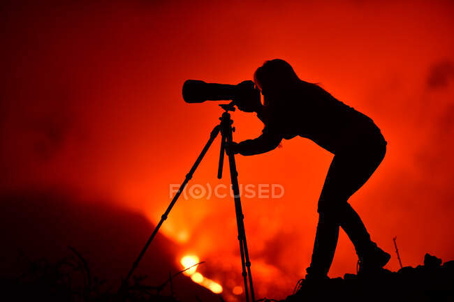 Vista laterale della silhouette di una donna accovacciata che fotografa con un teleobiettivo e treppiede l'esplosione di lava a La Palma Isole Canarie 2021 — Foto stock