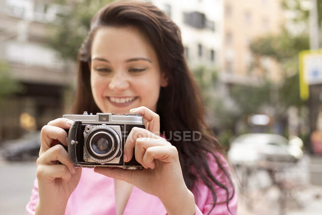 Joven mujer feliz con el pelo largo y castaño tomando fotos en la vieja cámara de fotos en la calle en la ciudad - foto de stock