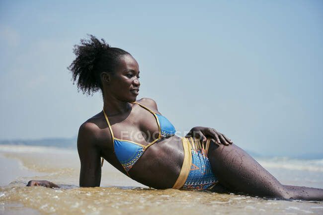 Joven y alegre hembra étnica en traje de baño con moño de pelo afro mirando hacia otro lado mientras yace en la costa del océano bajo el cielo azul - foto de stock