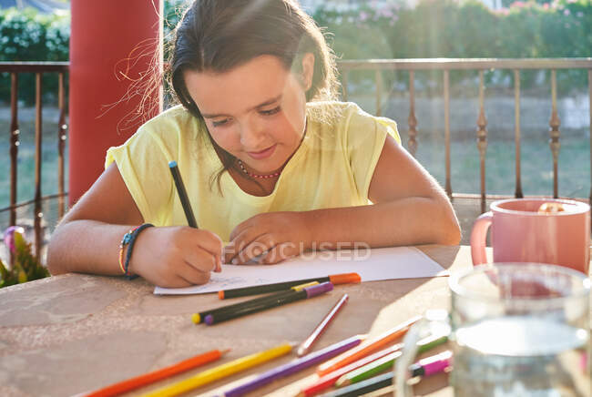 Chica positiva con el pelo oscuro en ropa casual sentado a la mesa con marcadores y dibujo sobre papel en la terraza en el día soleado - foto de stock