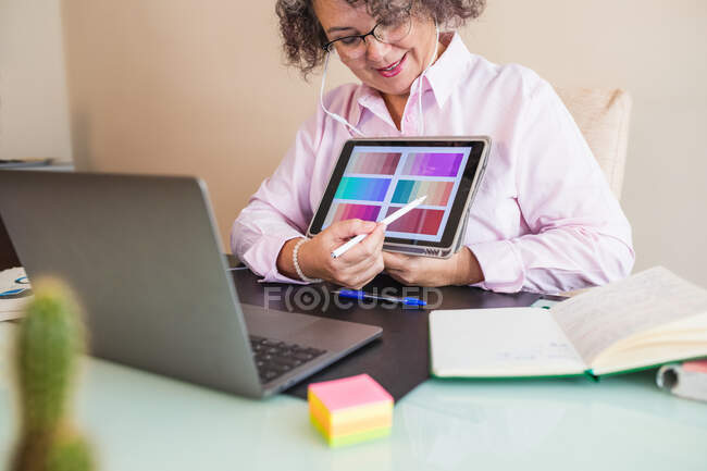 Crop senior female in earphones touching screen on tablet while pointing at color palette and speaking during video chat on netbook in office — Stock Photo