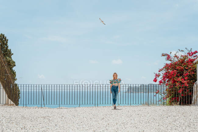 Повне тіло молодої жінки в модних джинсах і зверху стоячи на даху будинку, розташованому на узбережжі блакитного моря — стокове фото