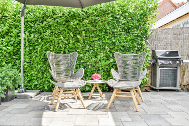 Armchairs and table with flowers in pot against gas grill and climbing plant on sunny day — Stock Photo