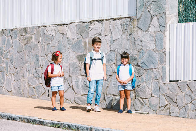 Écolier avec sac à dos parlant avec des amies debout sur un trottoir carrelé contre un mur de pierre au soleil — Photo de stock