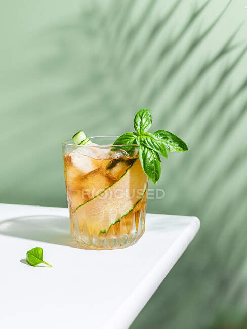 Bicchiere di cocktail freddo con fetta di cetriolo e foglie di basilico servito su tavolo bianco su sfondo verde — Foto stock
