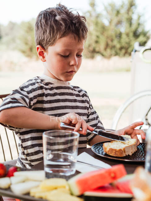Criança adorável focada manchando manteiga em fatia de pão enquanto toma café da manhã à mesa no pátio no verão — Fotografia de Stock