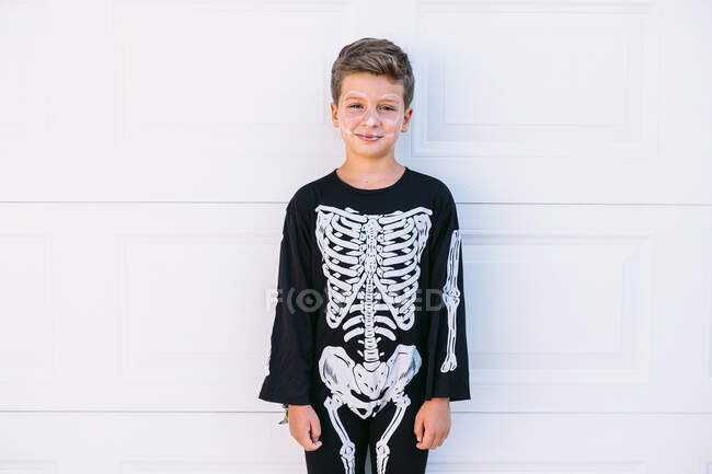 Хлопчик з білим пофарбованим скелетом, одягнений у чорний костюм на Хелловін, дивиться на камеру на білій стіні — стокове фото