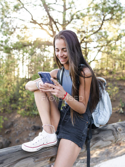 Cheerful cool female teenager with rucksack text messaging on cellphone while sitting on fence in Tenerife Spain — Stock Photo