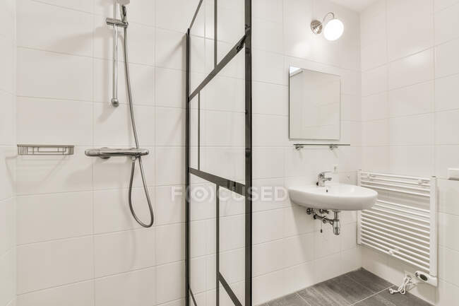 Минималистский дизайн белой ванной комнаты с раковиной под зеркалом, висящей на черепичной стене рядом со стеклянной душевой кабиной в квартире — стоковое фото