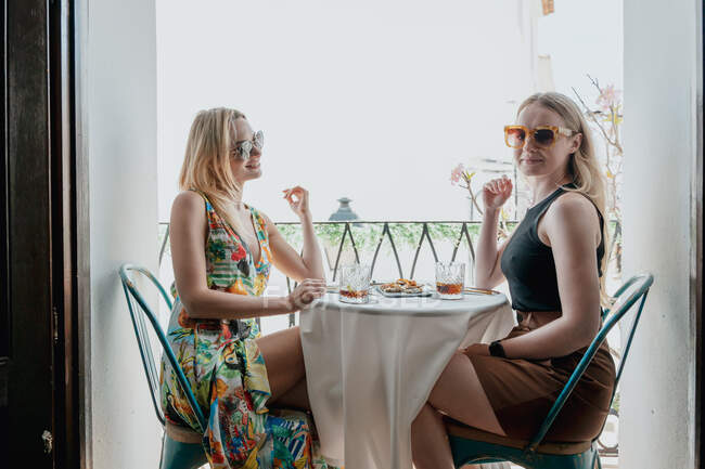 Vista lateral de amigas felizes almoçando à mesa e copos de coquetéis na varanda — Fotografia de Stock