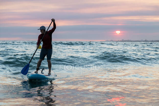Surfista masculino de fato de mergulho e chapéu na prancha de remo surfando na praia durante o pôr do sol — Fotografia de Stock