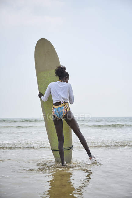 Rückenansicht einer afroamerikanischen Sportlerin mit Surfbrett, die das Meer vom sandigen Ufer aus unter wolkenlosem blauen Himmel bewundert — Stockfoto