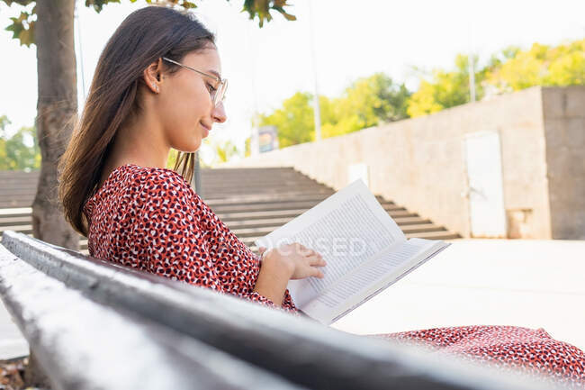 Jeune femme positive dans des vêtements élégants assis avec livre ouvert sur un banc en bois contre le bâtiment avec mur léger dans la journée — Photo de stock