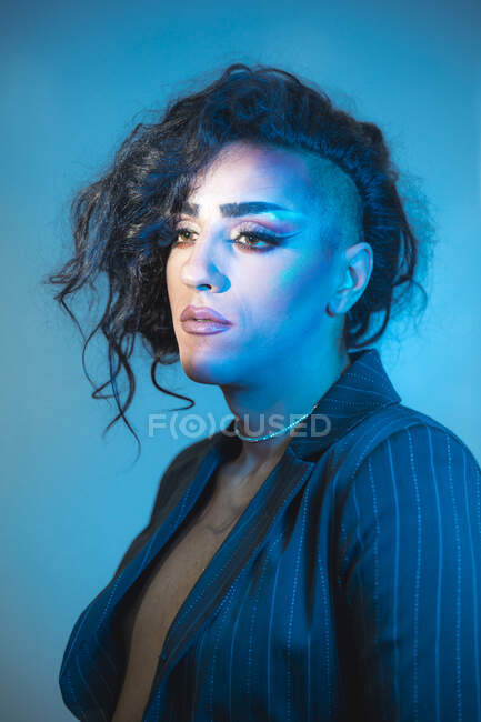 Jovem transexual modelo masculino com maquiagem na jaqueta elegante olhando para a câmera no fundo azul — Fotografia de Stock