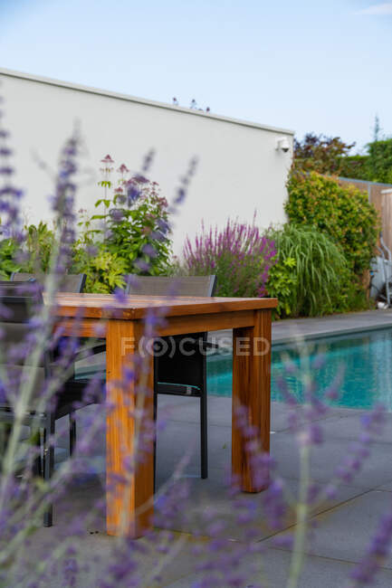 Table et chaises en bois placées près de la piscine sur une terrasse moderne décorée de plantes vertes en journée — Photo de stock
