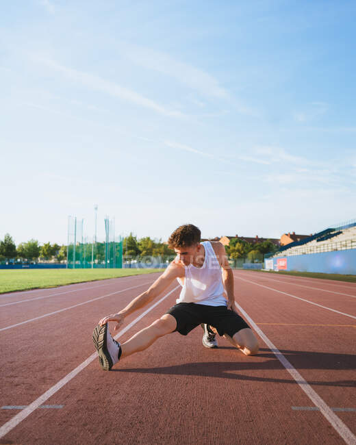 Adatta atleta di sesso maschile in abbigliamento sportivo appoggiato in avanti mentre accovacciato durante l'allenamento in pista sotto il cielo nuvoloso alla luce del sole — Foto stock