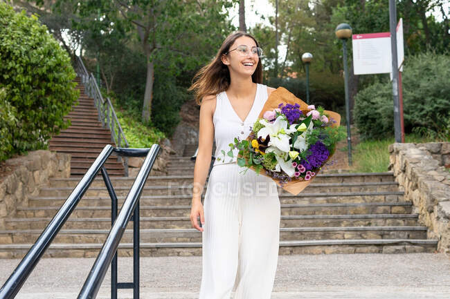 Zufriedene junge Frau mit Brille schaut weg, steht mit blühendem Blumenstrauß auf städtischen Treppen — Stockfoto