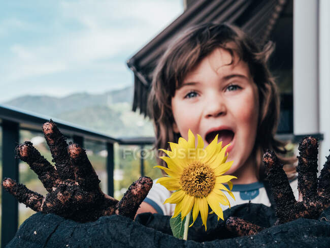 Enfant insouciant avec les mains sales et la bouche ouverte regardant vers l'avant contre le tournesol en fleurs sur le balcon dans la campagne — Photo de stock