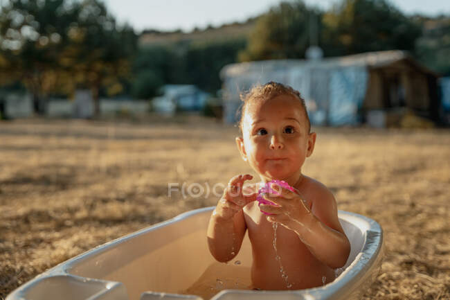 Niño pequeño con juguete sentado en un baño de plástico mientras juega con agua en el campo - foto de stock