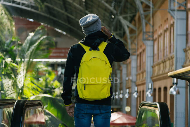 Back view of anonymous male hipster with bright yellow backpack standing near escalator while speaking on smartphone — Stock Photo
