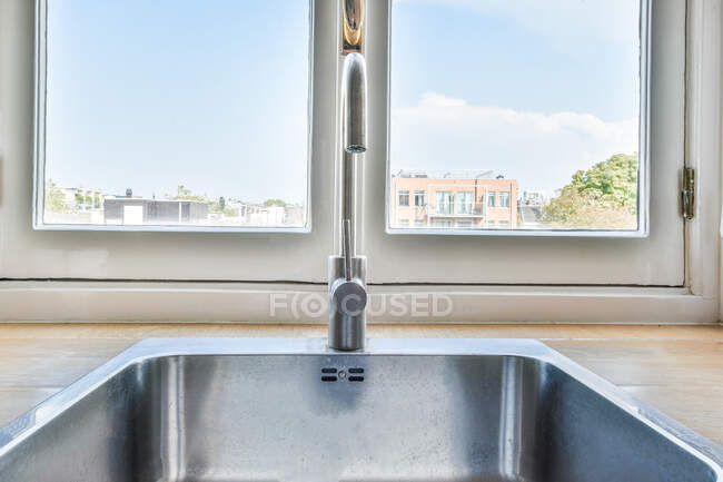 Comptoir en bois avec robinet chromé placé près de la fenêtre donnant sur la ville sous le ciel bleu en plein jour — Photo de stock