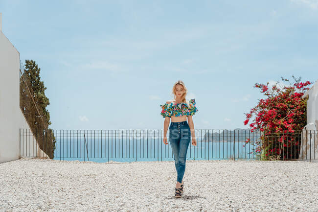 Full body of young female wearing trendy jeans and top standing on roof of house located on coast of blue sea — Stock Photo