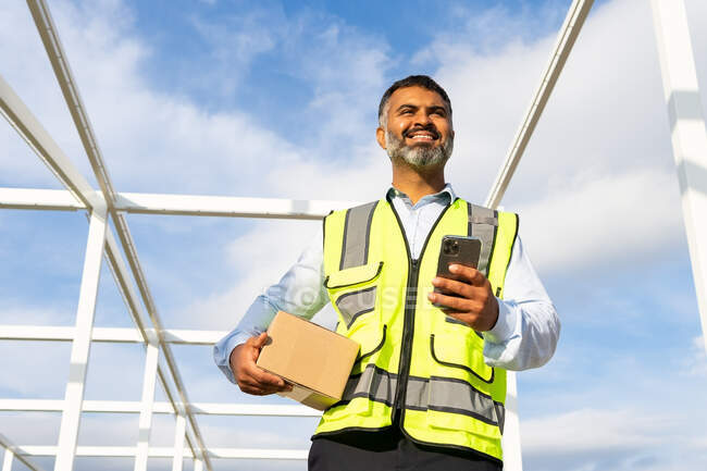 De baixo agradável barbudo trabalhador masculino étnico em uniforme pacote de transporte ao usar o telefone móvel no trabalho — Fotografia de Stock
