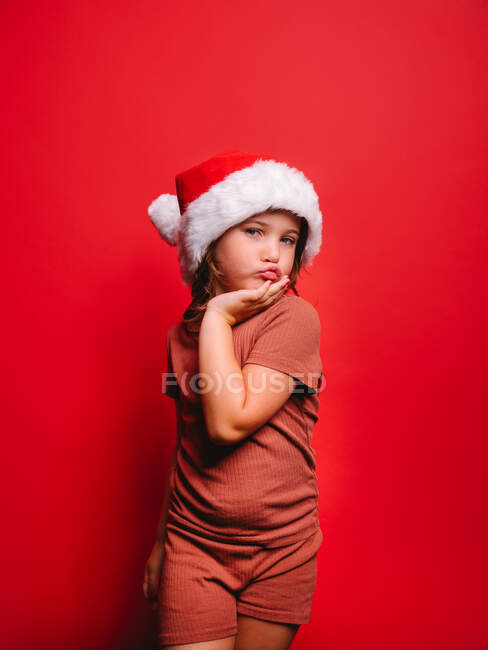 Adorable little girl in casual clothes and Santa hat pouting lips and touching face while standing against red background and looking at camera — Stock Photo