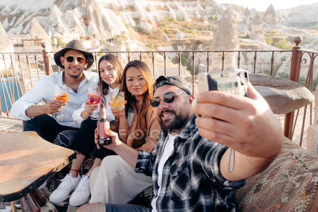 Male and female friends with cocktails and beer sitting on terrace of bar and taking self portrait on instant photo camera in terrace in Cappadocia, Turkey — Stock Photo