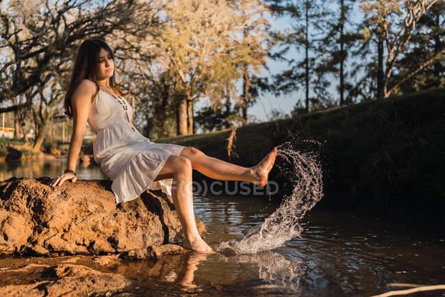 Charming female teenager in sundress having fun with splattering water while sitting on stone against river in park — Stock Photo