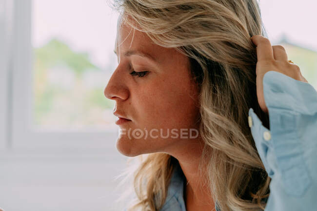 Seitenansicht erwachsene Frau mit welligen blonden Haaren, die Haare berühren, während sie im Haus auf dem Bett sitzt — Stockfoto