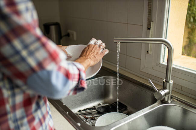 Вид збоку обрізаного невідомого чоловіка, що миє брудні тарілки, стоячи біля раковини на кухні та роблячи домашнє завдання — стокове фото