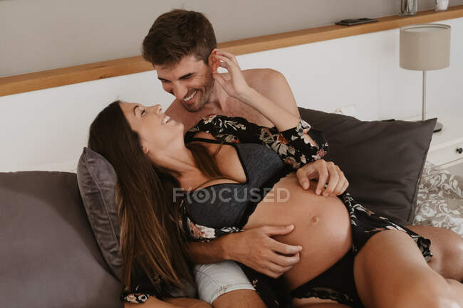 Zufriedener erwachsener Mann umarmt Bauch einer schwangeren Geliebten in Dessous, während er sich im Bett ansieht — Stockfoto