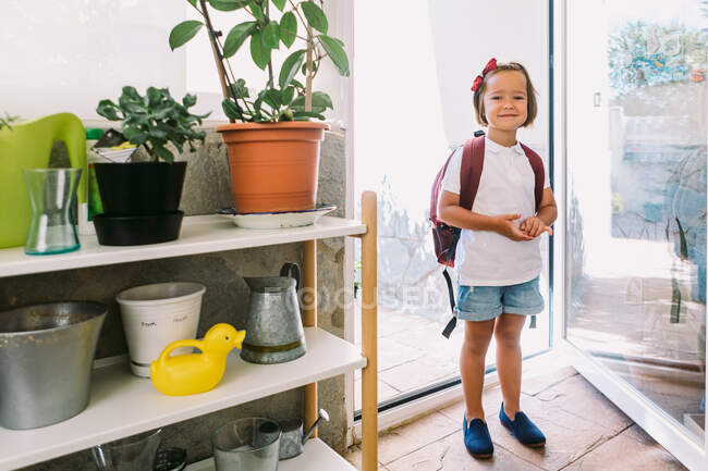 Écolière souriante avec sac à dos et arc sur les cheveux regardant la caméra entre porte en verre et plantes en pot à la maison — Photo de stock