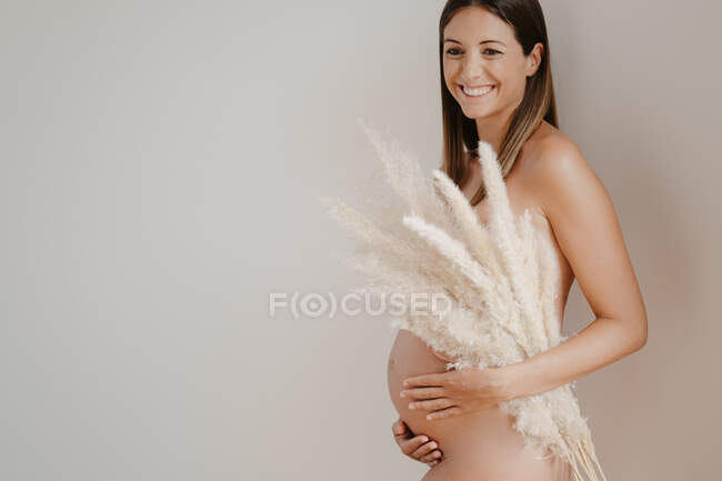 Side view of nude adult expectant female with soft plant sprigs caressing belly while looking away on light background — Stock Photo