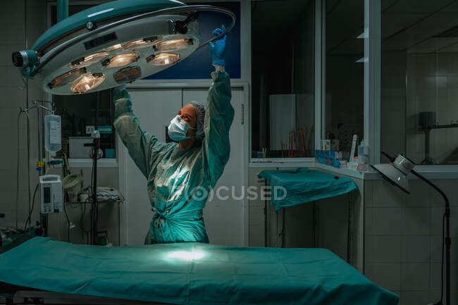 Joven veterinaria en uniforme quirúrgico y máscara estéril mirando hacia arriba mientras ajusta la lámpara por encima del textil médico en la clínica - foto de stock